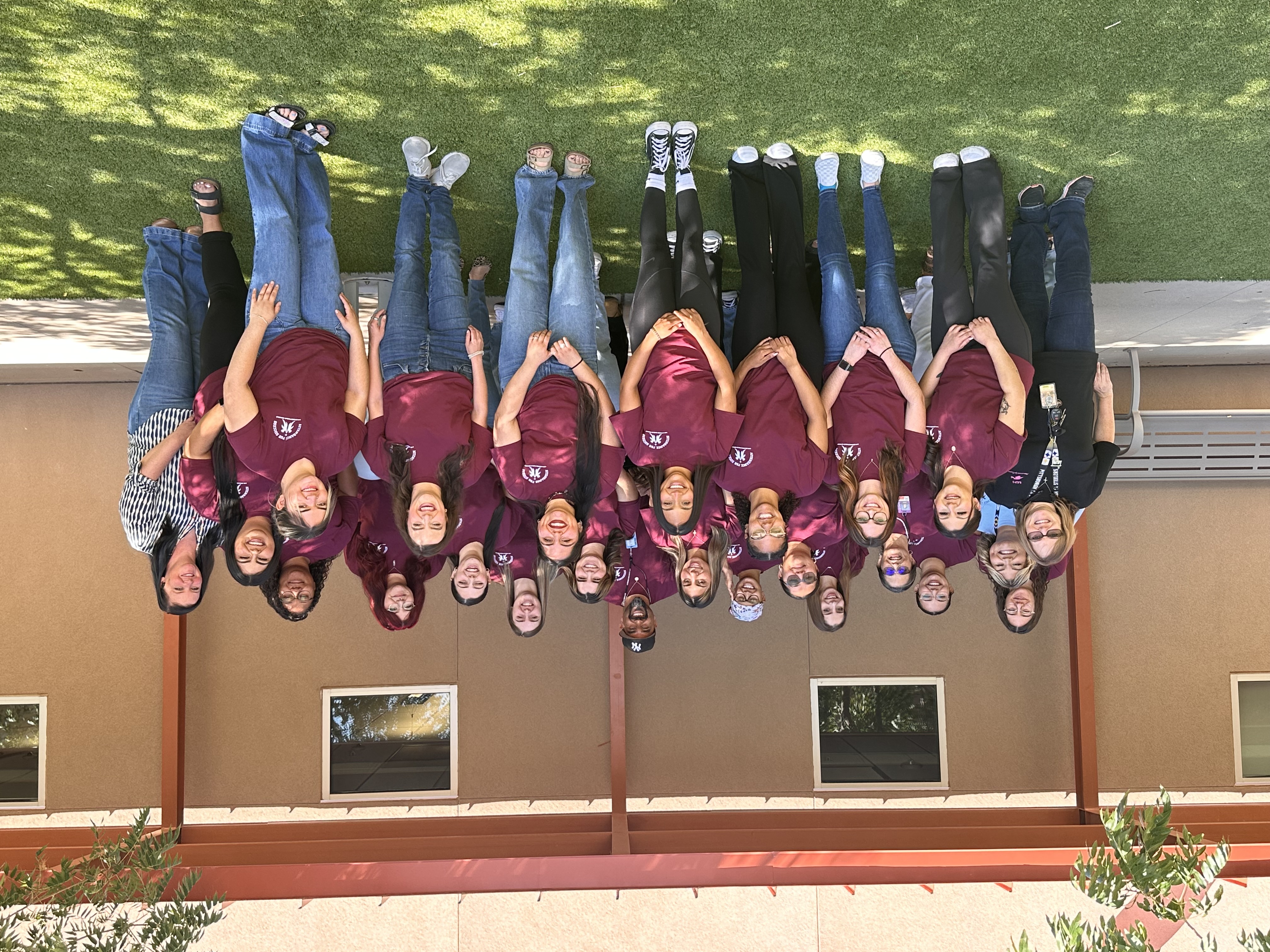 Group photo of a diverse team wearing matching maroon shirts, smiling outdoors in front of a building.