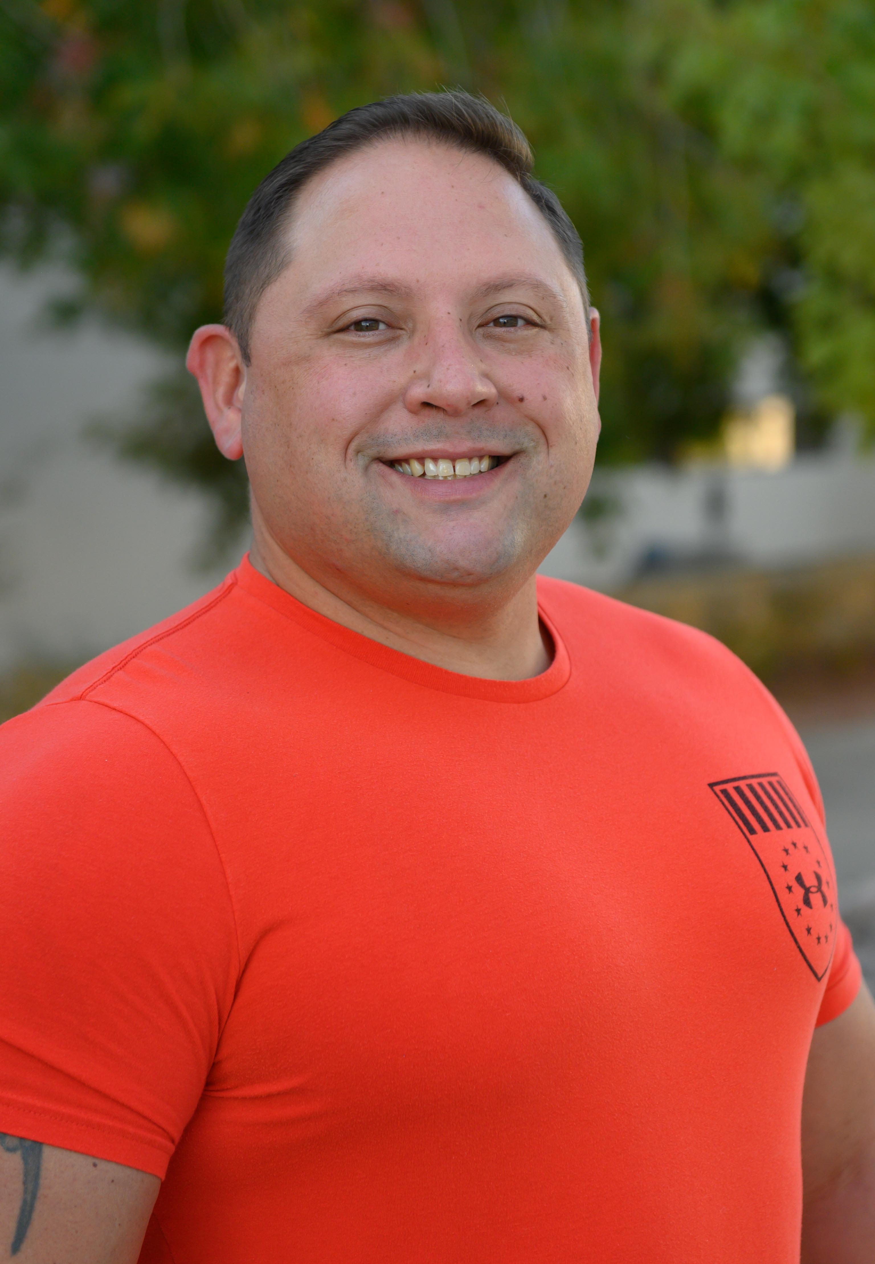 A smiling man wearing an orange shirt stands outdoors with trees in the background.