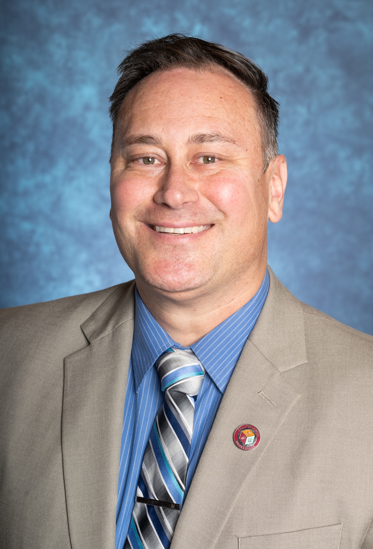 Portrait of a professional man in a suit with a blue shirt and striped tie, smiling against a blue textured background.