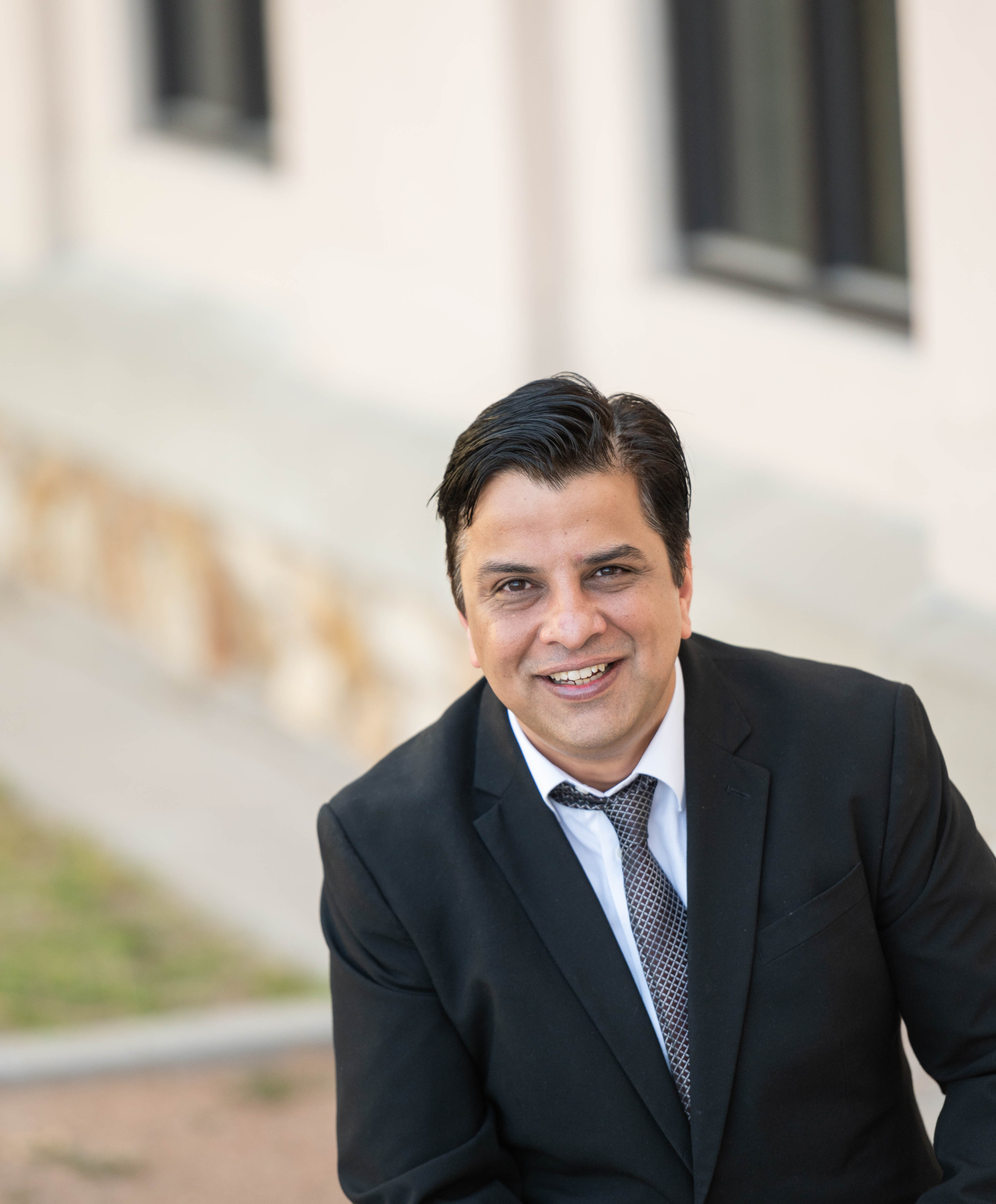 A smiling man in a black suit and tie poses confidently outdoors, with a blurred building in the background.