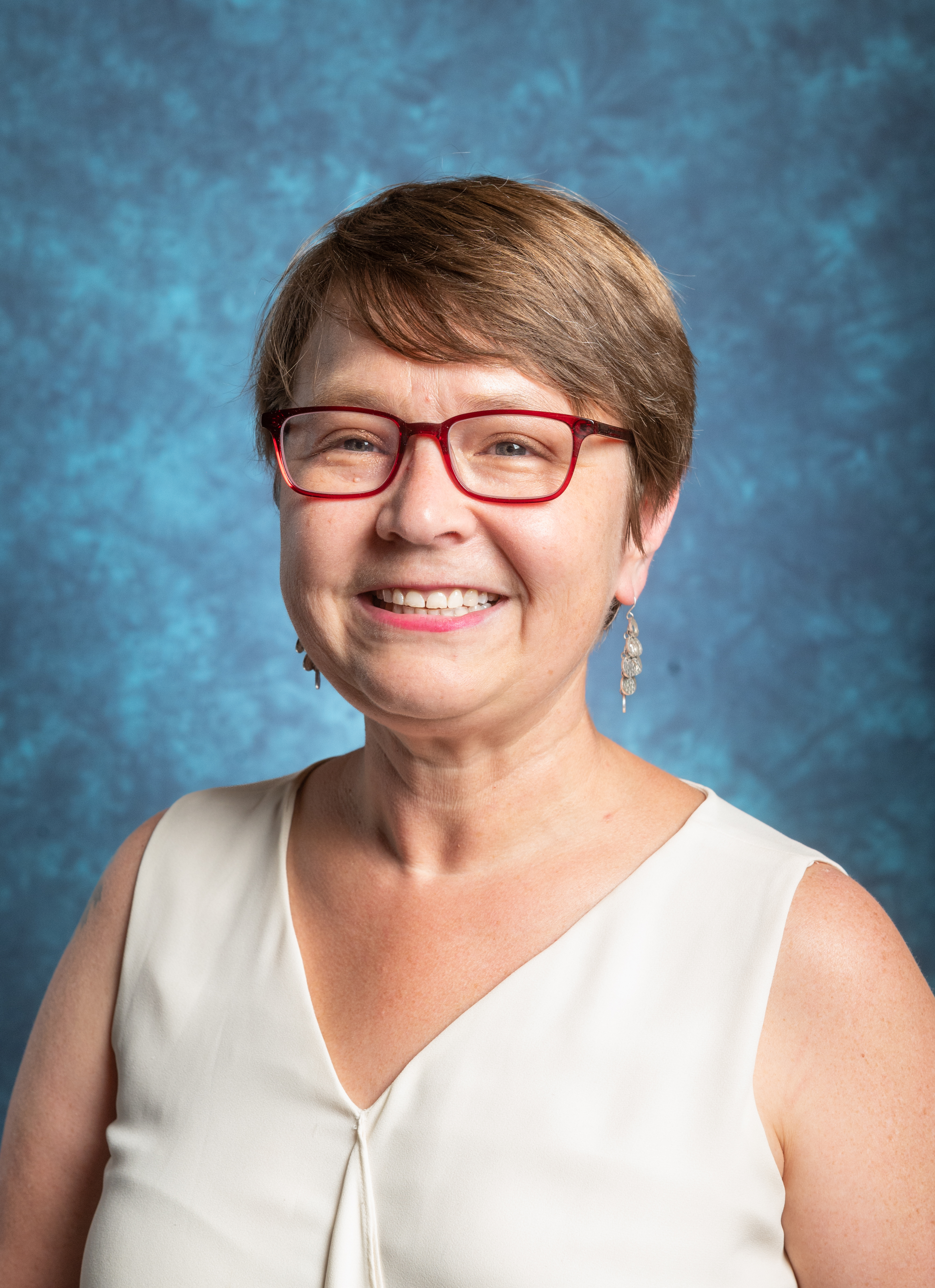 A smiling woman with short hair and red glasses, wearing a sleeveless white top, against a textured blue background.