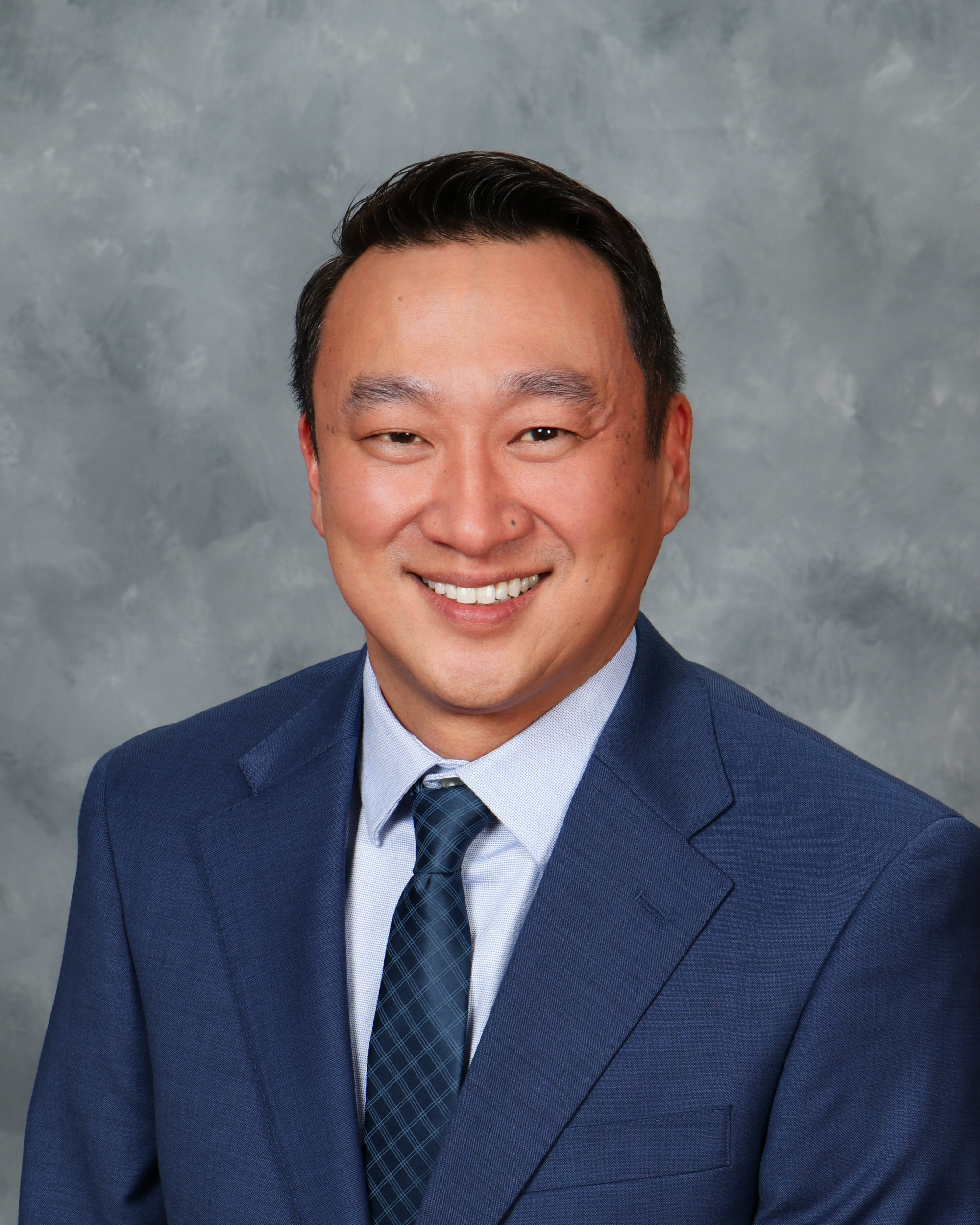 Professional headshot of a smiling man in a blue suit and tie, set against a gray background.